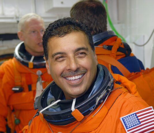 José Hernández Joins NASA’s Discovery Mission Astronaut José Hernández, a Hispanic man, smiles broadly at the camera while waiting to board space shuttle Discovery. He wears an orange escape suit that has an American flag patch on the left shoulder. Behind him are mission specialists Patrick Forrester (left) and Christer Fuglesang (back to camera).