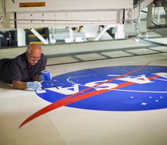 NASA Celebrates 65 Years of Iconic Logo, Media Invited Image of a man on the ground painting blue on the NASA meatball logo.