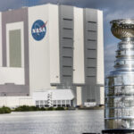 Stanley Cup Visits NASA’s Kennedy Space Center The silver Stanley Cup is in the foreground at right. The Stanley Cup looks like a large silver bowl set on top of a stacked column of thick silver layers. There are words engraved on the Cup. Behind it is a body of water and the Vehicle Assembly Building at NASA