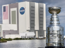 Stanley Cup Visits NASA’s Kennedy Space Center The silver Stanley Cup is in the foreground at right. The Stanley Cup looks like a large silver bowl set on top of a stacked column of thick silver layers. There are words engraved on the Cup. Behind it is a body of water and the Vehicle Assembly Building at NASA