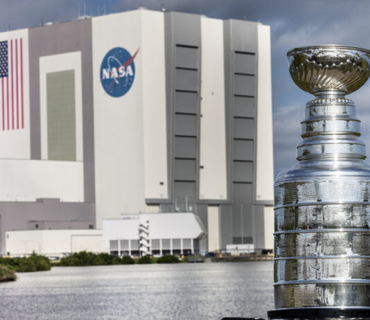 Stanley Cup Visits NASA’s Kennedy Space Center The silver Stanley Cup is in the foreground at right. The Stanley Cup looks like a large silver bowl set on top of a stacked column of thick silver layers. There are words engraved on the Cup. Behind it is a body of water and the Vehicle Assembly Building at NASA