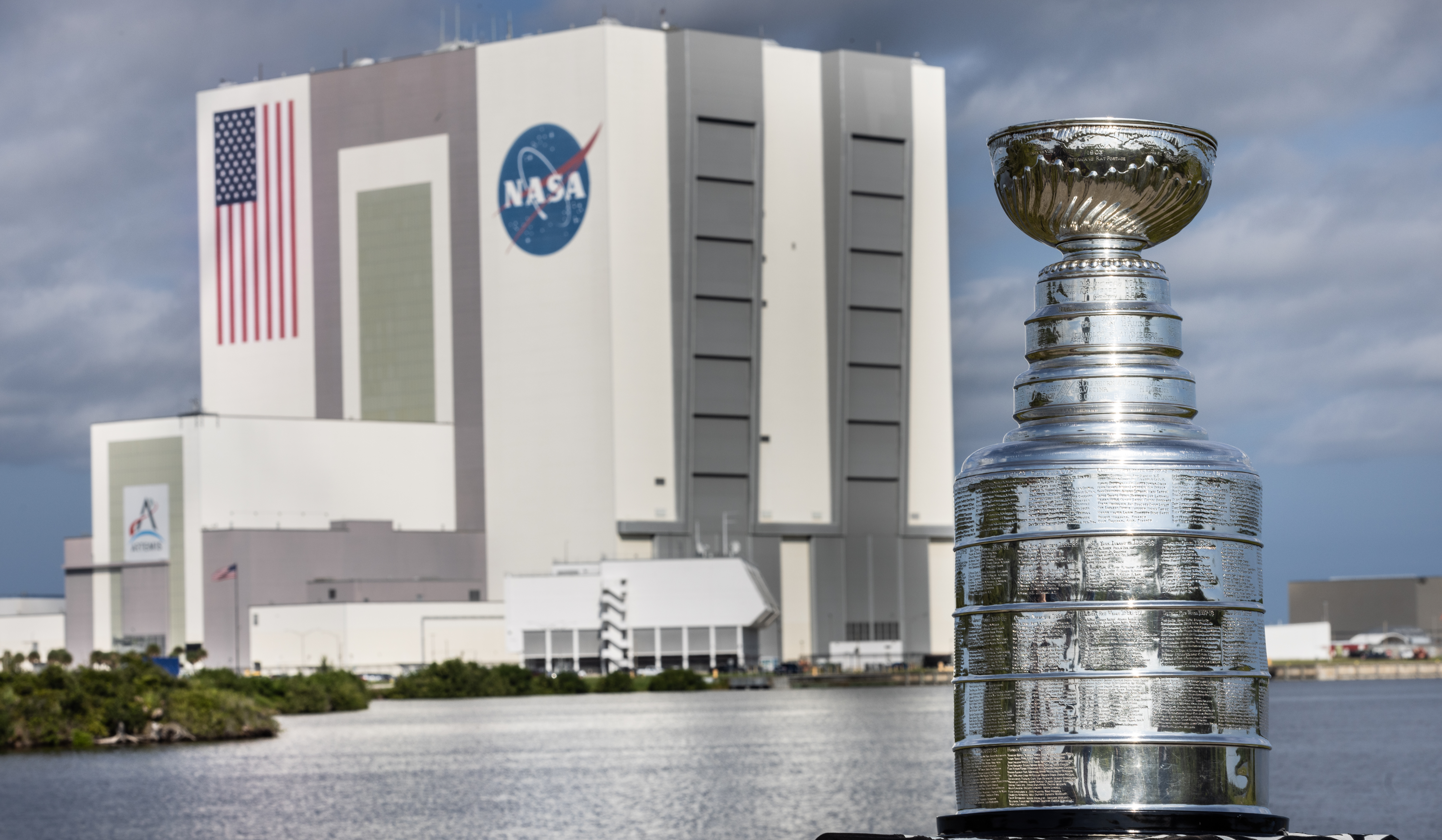 The silver Stanley Cup is in the foreground at right. The Stanley Cup looks like a large silver bowl set on top of a stacked column of thick silver layers. There are words engraved on the Cup. Behind it is a body of water and the Vehicle Assembly Building at NASA