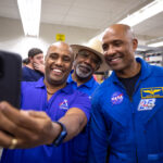 Astronaut Victor Glover Hosts Selfie Session with NASA Fans Three African American men - two Kennedy Space Center employees wearing blue polo shirts with an Artemis logo (left, middle back) and NASA astronaut Victor Glover (right) - pose for a photo taken with a cell phone.