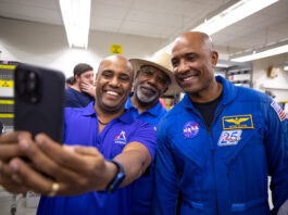 Astronaut Victor Glover Hosts Selfie Session with NASA Fans Three African American men - two Kennedy Space Center employees wearing blue polo shirts with an Artemis logo (left, middle back) and NASA astronaut Victor Glover (right) - pose for a photo taken with a cell phone.