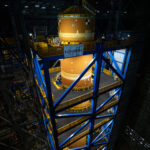 Artemis II Core Stage Relocated to High Bay 2 An immense orange cylindrical object - the SLS core stage - is surrounded by a blue metal structure. The space around them is dark.