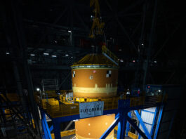 Artemis II Core Stage Relocated to High Bay 2 An immense orange cylindrical object - the SLS core stage - is surrounded by a blue metal structure. The space around them is dark.