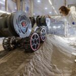 NASA Kennedy Conducts Newest Robot Experiment Image shows a robot on a mound of simulated lunar soil with a man in a white bunny suit observing
