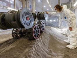 NASA Kennedy Conducts Newest Robot Experiment Image shows a robot on a mound of simulated lunar soil with a man in a white bunny suit observing