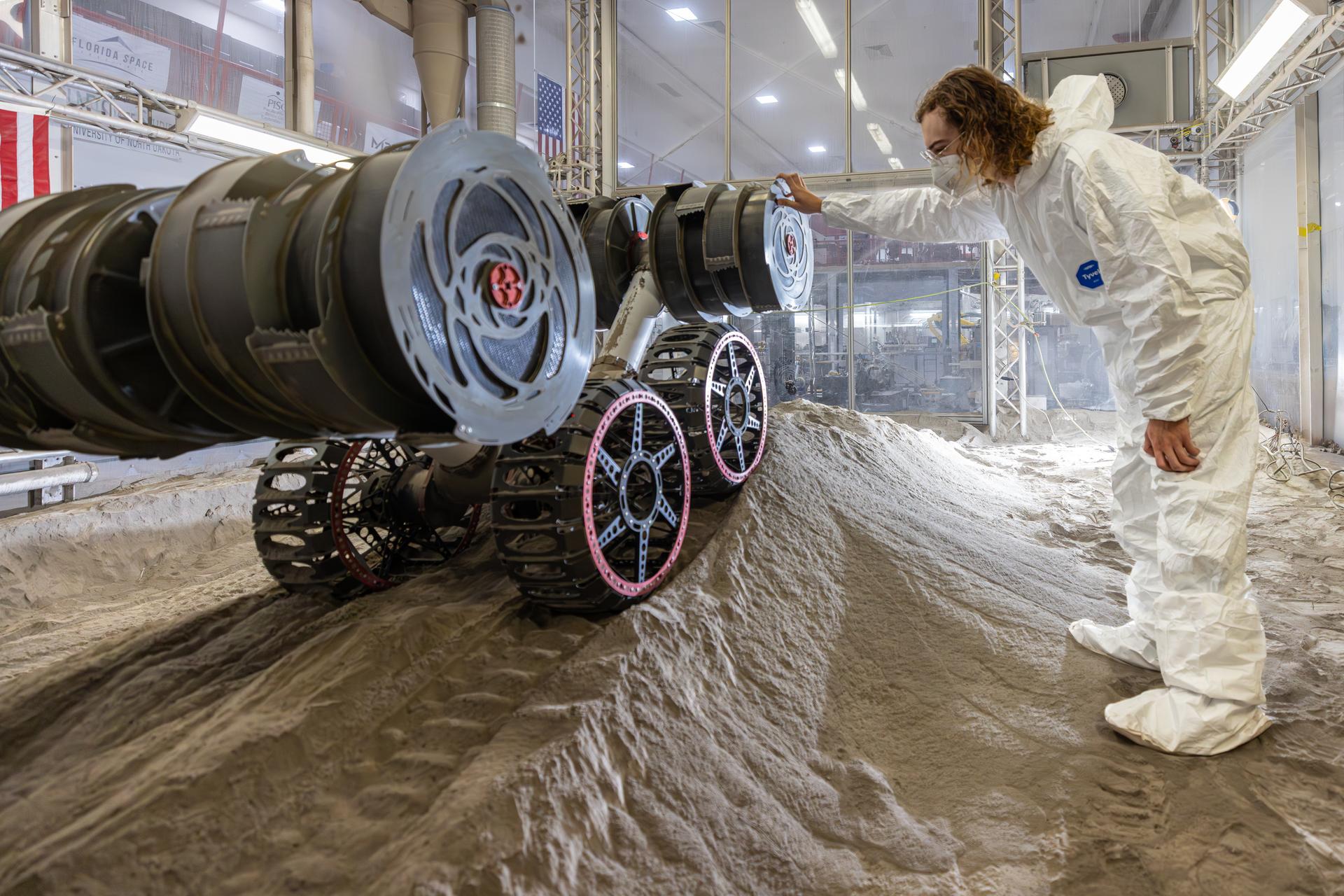 Image shows a robot on a mound of simulated lunar soil with a man in a white bunny suit observing
