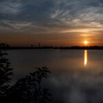 Crew-11 Launch Attempt at Dawn by NASA In the distance, the Sun rises, casting an orange glow over the surrounding sky. It is also reflected in the water below it, where it looks like a column of orange light. The outlines of several structures are visible in the middle ground. At the top of the photo are white clouds that look like streaks of paint. At the bottom of the photo, those clouds are partially reflected in the water (at right) and some foliage peeks in (at left).