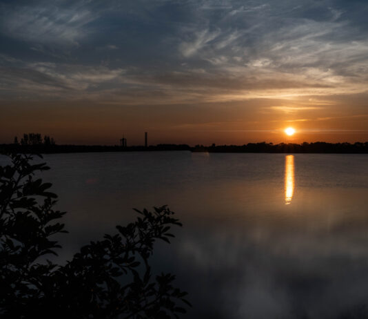 Crew-11 Launch Attempt at Dawn by NASA In the distance, the Sun rises, casting an orange glow over the surrounding sky. It is also reflected in the water below it, where it looks like a column of orange light. The outlines of several structures are visible in the middle ground. At the top of the photo are white clouds that look like streaks of paint. At the bottom of the photo, those clouds are partially reflected in the water (at right) and some foliage peeks in (at left).