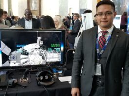Lee Bingham Heads Lunar Simulation Support for NASA Artemis Mission NASA employee Lee Bingham, wearing a dark suit with a vest and red tie, stands next to a computer monitor with a Gateway simulation displayed on the screen.