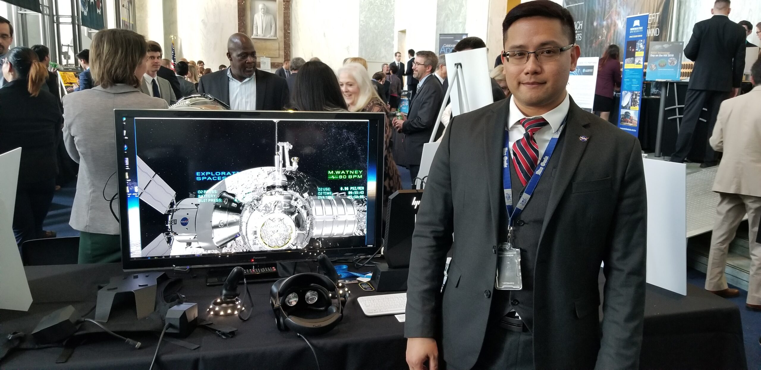 lee bingham demoing a gateway eva simulator in washington dc at nasas tech day on the hill.jpg NASA employee Lee Bingham, wearing a dark suit with a vest and red tie, stands next to a computer monitor with a Gateway simulation displayed on the screen.