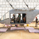 NASA’s Drop Test Advances Safety in Air Taxi Innovations A white aircraft body with small black dots is hoisted in the air by cables. There are several test dummies inside of the aircraft. The aircraft does not hang far off of the ground. Three workers with hardhats remove pink foam squares from underneath the aircraft. Behind the aircraft is a large white board to measure the height, which includes many black squares with small numbers inside some of the bottom squares.