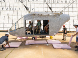 NASA’s Drop Test Advances Safety in Air Taxi Innovations A white aircraft body with small black dots is hoisted in the air by cables. There are several test dummies inside of the aircraft. The aircraft does not hang far off of the ground. Three workers with hardhats remove pink foam squares from underneath the aircraft. Behind the aircraft is a large white board to measure the height, which includes many black squares with small numbers inside some of the bottom squares.