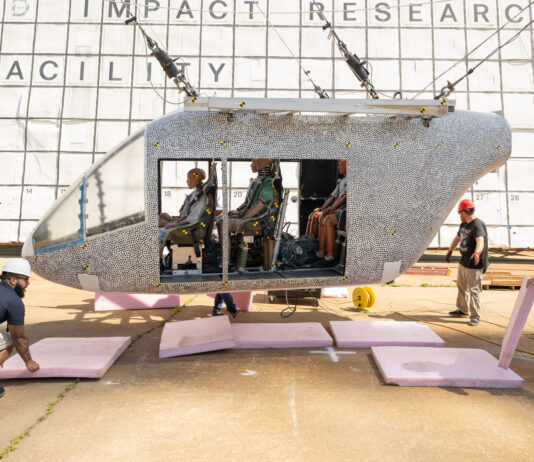 NASA’s Drop Test Advances Safety in Air Taxi Innovations A white aircraft body with small black dots is hoisted in the air by cables. There are several test dummies inside of the aircraft. The aircraft does not hang far off of the ground. Three workers with hardhats remove pink foam squares from underneath the aircraft. Behind the aircraft is a large white board to measure the height, which includes many black squares with small numbers inside some of the bottom squares.