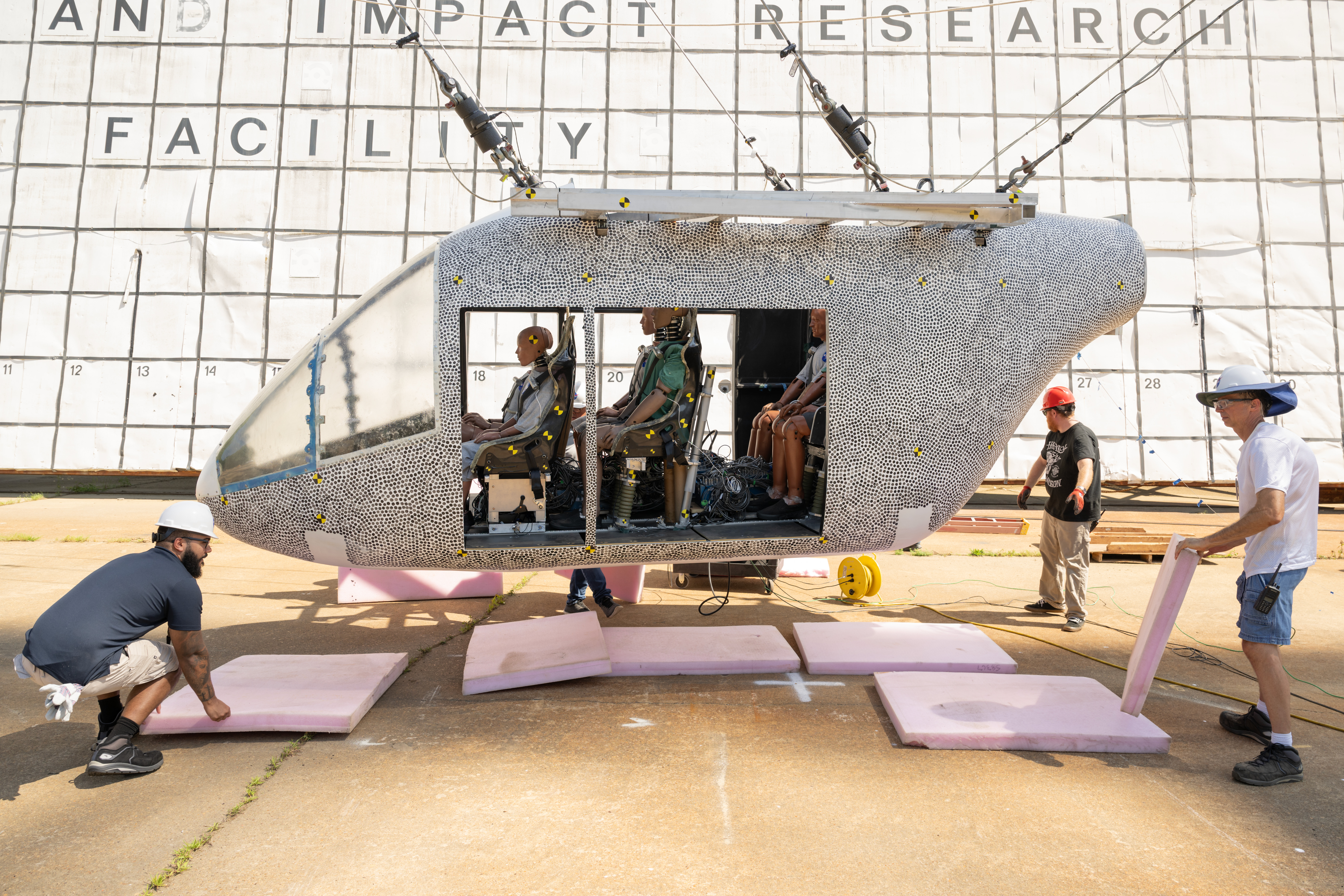 lrc 2025 ocio p 01814orig.jpg A white aircraft body with small black dots is hoisted in the air by cables. There are several test dummies inside of the aircraft. The aircraft does not hang far off of the ground. Three workers with hardhats remove pink foam squares from underneath the aircraft. Behind the aircraft is a large white board to measure the height, which includes many black squares with small numbers inside some of the bottom squares.