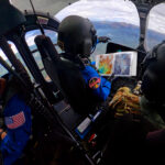 NASA Initiates Helicopter Training for Artemis Space Missions NASA astronauts Matthew Dominick (left, in a blue jumpsuit with a patch on the right arm) and Mark Vande Hei (right, in camouflage clothing) in the cockpit of a helicopter. Dominick holds an open binder in his right hand and points forward with his left. They are both wearing black helmets. There is another person behind Dominick, also wearing a blue jumpsuit and helmet, facing the opposite way.