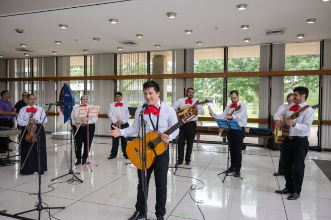 manuel retana singing with jsc mariachi celestial in 2024 diversity day.jpg A group of musicians, dressed in traditional mariachi attire with white shirts, red bow ties, and black pants, perform in a bright, spacious room. The lead musician stands at the front, holding a guitar and singing into a microphone.