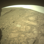 Challenging NASA Experiment Conducted at Witch Hazel Hill A color image from the Martian surface shows uneven yellow-tan ground, covered in gravel and much larger flat, angular rocks showing above the surface. Parts of the rover are visible but shaded – a portion across the top of the frame, and a wheel in the lower left corner. A drill hole is visible in the ground near the center of the image, surrounded by lighter-colored soil.