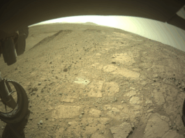 Challenging NASA Experiment Conducted at Witch Hazel Hill A color image from the Martian surface shows uneven yellow-tan ground, covered in gravel and much larger flat, angular rocks showing above the surface. Parts of the rover are visible but shaded – a portion across the top of the frame, and a wheel in the lower left corner. A drill hole is visible in the ground near the center of the image, surrounded by lighter-colored soil.