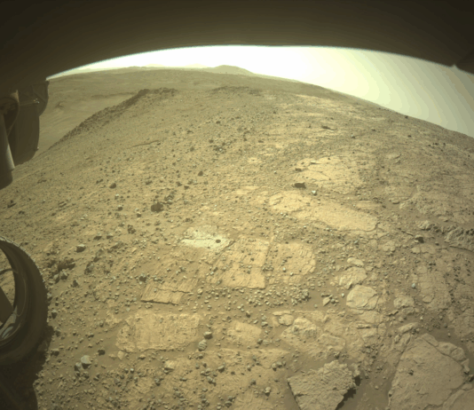 Challenging NASA Experiment Conducted at Witch Hazel Hill A color image from the Martian surface shows uneven yellow-tan ground, covered in gravel and much larger flat, angular rocks showing above the surface. Parts of the rover are visible but shaded – a portion across the top of the frame, and a wheel in the lower left corner. A drill hole is visible in the ground near the center of the image, surrounded by lighter-colored soil.