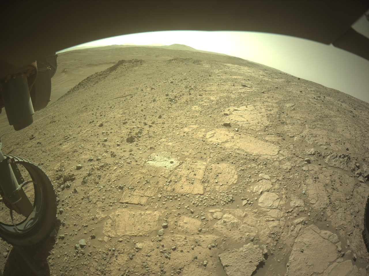 mars perseverance flf 1497 0799847065 176ecm n0730000fhaz00219 01 295j.png A color image from the Martian surface shows uneven yellow-tan ground, covered in gravel and much larger flat, angular rocks showing above the surface. Parts of the rover are visible but shaded – a portion across the top of the frame, and a wheel in the lower left corner. A drill hole is visible in the ground near the center of the image, surrounded by lighter-colored soil.