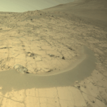 NASA Hunts for Primeval Rocks in Forlandet Flats A color image from the Martian surface shows uneven yellow-tan ground that rises from the foreground upward toward the upper right corner of the image and off the frame. The ground appears dry, with narrow cracks dividing the ground into squares and other angular shapes. A narrow strip of smoother ground, like a low berm of mounded sand, curves from the left side toward the middle of the image. Beyond that, toward the upper left, the dry ground is more gravel-covered.