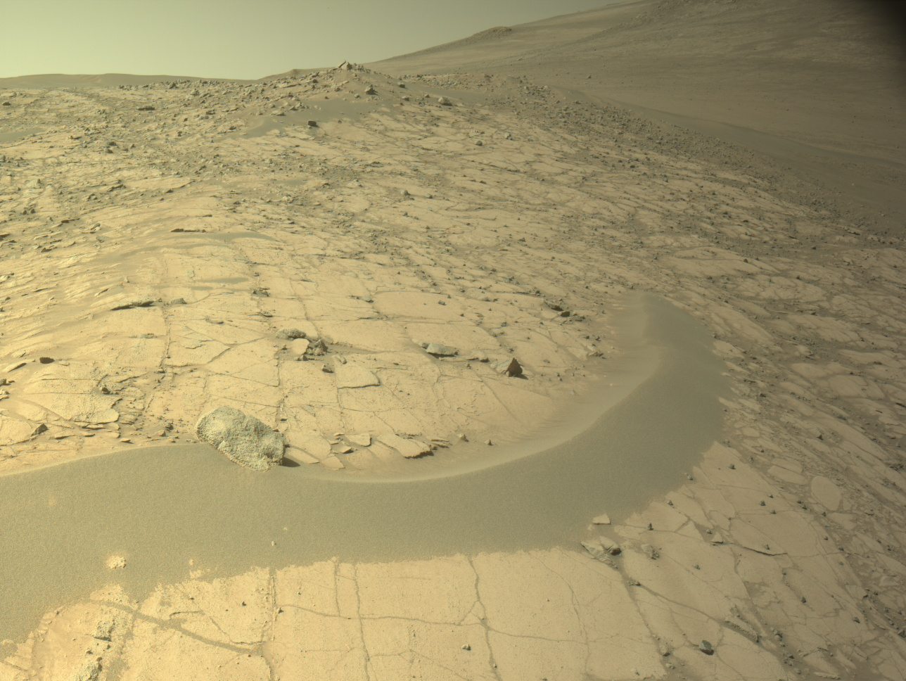 mars perseverance nlf 1512 0801175461 522ecm n0740000ncam02512 04 195j.png A color image from the Martian surface shows uneven yellow-tan ground that rises from the foreground upward toward the upper right corner of the image and off the frame. The ground appears dry, with narrow cracks dividing the ground into squares and other angular shapes. A narrow strip of smoother ground, like a low berm of mounded sand, curves from the left side toward the middle of the image. Beyond that, toward the upper left, the dry ground is more gravel-covered.