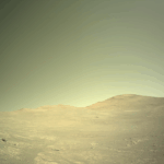 NASA’s Perseverance Rover Encounters Megabreccia on Mars A color photo from the Martian surface shows a landscape with terrain appearing light mustard brown, beneath a sky of gray-green gold. The ground is hilly but fairly smooth, with a few small scattered rocks, and a wavy horizon line about mid-frame.