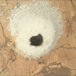 NASA’s Flattening Experiment: A Scientific Triumph A color photograph from the Martian surface shows a close-up, overhead view of flat, cracked terrain colored pale orange-tan. At the center of the image a circular hole has been dug into the ground, shadowed and dark, and it’s surrounded by mounded grainy soil, colored a very light golden tan, that appears to have been dug out of the hole.