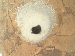 NASA’s Flattening Experiment: A Scientific Triumph A color photograph from the Martian surface shows a close-up, overhead view of flat, cracked terrain colored pale orange-tan. At the center of the image a circular hole has been dug into the ground, shadowed and dark, and it’s surrounded by mounded grainy soil, colored a very light golden tan, that appears to have been dug out of the hole.