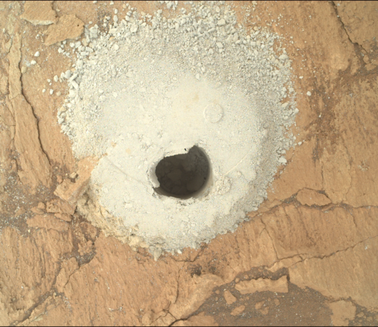 NASA’s Flattening Experiment: A Scientific Triumph A color photograph from the Martian surface shows a close-up, overhead view of flat, cracked terrain colored pale orange-tan. At the center of the image a circular hole has been dug into the ground, shadowed and dark, and it’s surrounded by mounded grainy soil, colored a very light golden tan, that appears to have been dug out of the hole.