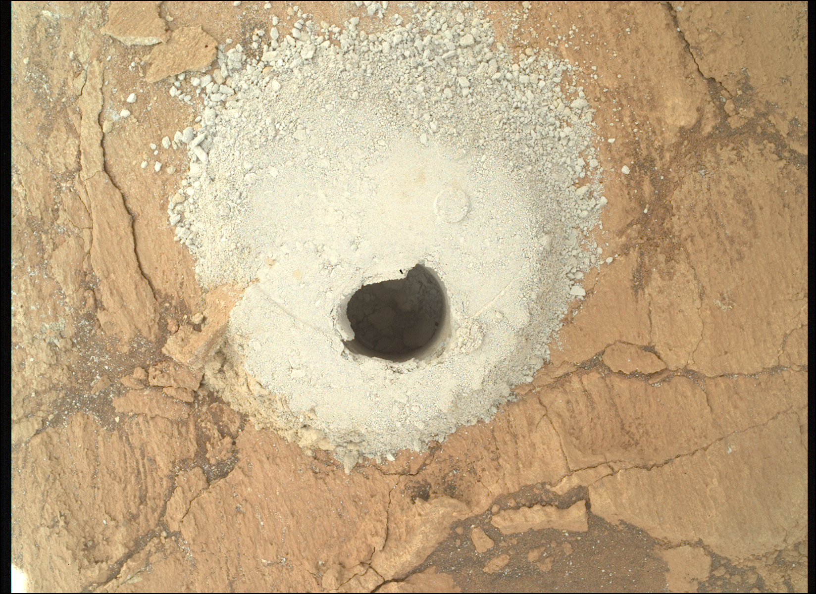 A color photograph from the Martian surface shows a close-up, overhead view of flat, cracked terrain colored pale orange-tan. At the center of the image a circular hole has been dug into the ground, shadowed and dark, and it’s surrounded by mounded grainy soil, colored a very light golden tan, that appears to have been dug out of the hole.