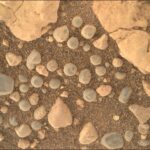 NASA’s Discovery: Mysterious Sand Spheres Uncovered A close-up color photograph from the Martian surface shows an overhead view of a patch of ground covered in what looks like very coarse, rust-colored sand, with numerous rocks in various colors resting atop the sand. Large pale-orange rocks, knobby and lined with small fractures, dominate the upper right and left corners of the frame. Most of the rest of the image is highlighted by dozens of smaller rocks, about half of them pale gray and nearly spherical, resembling dust-covered blueberries or frozen peas.