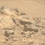 NASA Explores Mars En Route to Distant Solar Regions A color photo from the Martian surface shows pale orange, very rocky terrain in the foreground, taking up the lower two-thirds of the frame, creating a horizon line from the upper left corner of the image to the middle right side. The terrain beyond, in the distance, is blurry and takes up the rest of the frame. The rocks in the foreground range from tiny, sharp stones to large outcroppings, the latter showing some medium gray highlights among the orange rock and dust that comprise them. The largest rocks, on the left side, show numerous grooves and cracks on their faces, ranging from vertical to slightly left-skewing.