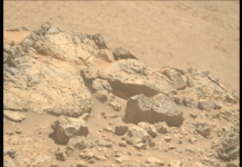 NASA Explores Mars En Route to Distant Solar Regions A color photo from the Martian surface shows pale orange, very rocky terrain in the foreground, taking up the lower two-thirds of the frame, creating a horizon line from the upper left corner of the image to the middle right side. The terrain beyond, in the distance, is blurry and takes up the rest of the frame. The rocks in the foreground range from tiny, sharp stones to large outcroppings, the latter showing some medium gray highlights among the orange rock and dust that comprise them. The largest rocks, on the left side, show numerous grooves and cracks on their faces, ranging from vertical to slightly left-skewing.