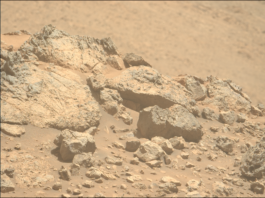 NASA Explores Mars En Route to Distant Solar Regions A color photo from the Martian surface shows pale orange, very rocky terrain in the foreground, taking up the lower two-thirds of the frame, creating a horizon line from the upper left corner of the image to the middle right side. The terrain beyond, in the distance, is blurry and takes up the rest of the frame. The rocks in the foreground range from tiny, sharp stones to large outcroppings, the latter showing some medium gray highlights among the orange rock and dust that comprise them. The largest rocks, on the left side, show numerous grooves and cracks on their faces, ranging from vertical to slightly left-skewing.