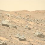 Perseverance Hunts for Signs of Contact Metamorphism on Mars A color photo from the surface of Mars shows very rocky terrain in the foreground sloping downward from left to right. The ground ranges in shades from orange-tan to brown, while the rocks are mostly gray. A trio of hills rises up in the distance.