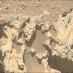 NASA Investigates Unidentified Object Approaching Earth A color photo from the Martian surface shows a close-up of a large tan and pale orange rock pockmarked with numerous large holes, which are mostly filled with dark, rust-colored sand. In the background, pale orange, very rocky terrain is visible in the top third of the image.