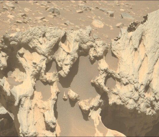 NASA Investigates Unidentified Object Approaching Earth A color photo from the Martian surface shows a close-up of a large tan and pale orange rock pockmarked with numerous large holes, which are mostly filled with dark, rust-colored sand. In the background, pale orange, very rocky terrain is visible in the top third of the image.