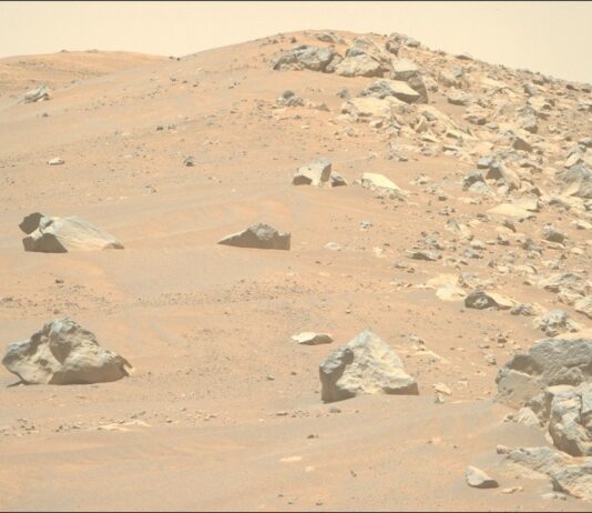 Connecting Regional Rock Formations to Broader Terrain: NASA Study A color photo from the Martian surface shows a landscape of sandy, rocky terrain. What looks like a pair of light orange-brown sand dunes rise in the background. Four or five large, sharp-edged rocks are strewn among the sand on the left side of the image. But a clear vertical dividing line extends from the center of the image to the right side, where the sand is covered by similar rocks of all sizes. A pale yellow sky is visible above the scene.