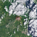 Mayon volcano erupts, NASA monitors lava flow. The upper slopes of Mayon volcano appear brown, with several narrow channels radiating from the crater. A red infrared heat signature appears near the summit, with red streaks extending east and southeast. The lower slopes are green and forested. Farmland and towns are visible in the lower part of the image.