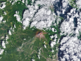 Mayon volcano erupts, NASA monitors lava flow. The upper slopes of Mayon volcano appear brown, with several narrow channels radiating from the crater. A red infrared heat signature appears near the summit, with red streaks extending east and southeast. The lower slopes are green and forested. Farmland and towns are visible in the lower part of the image.