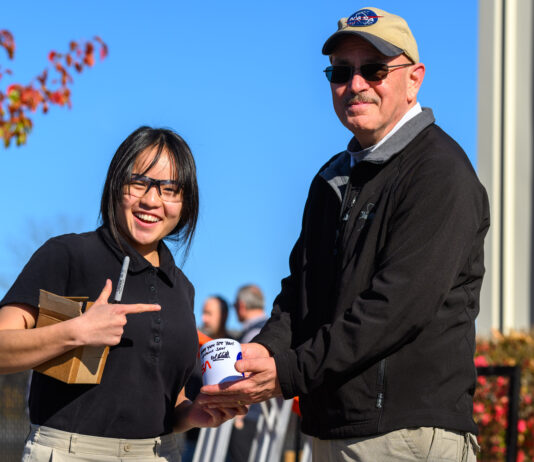 NASA and Notre Dame Unite to Encourage STEM Career Aspirations Portage School of Leaders High School junior Rebecca Anderson, wearing a black short-sleeved shirt and glasses, stands smiling beside NASA Glenn’s Daniel Sutliff, an acoustic engineer, on the campus of the University of Notre Dame. Sutliff is wearing a NASA ballcap and black jacket. He and Anderson are holding an autographed NASA mug, which Sutliff signed with a black ink permanent magic marker. Anderson is holding the black marker and a small cardboard box.