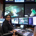 From Oceans to Space: Aaisha Ali’s NASA Journey A woman sits at a desk and poses in front of three monitors. Multiple monitors display live data and video of astronauts on the International Space Station. An American flag and NASA mission patches are visible on the wall behind her.