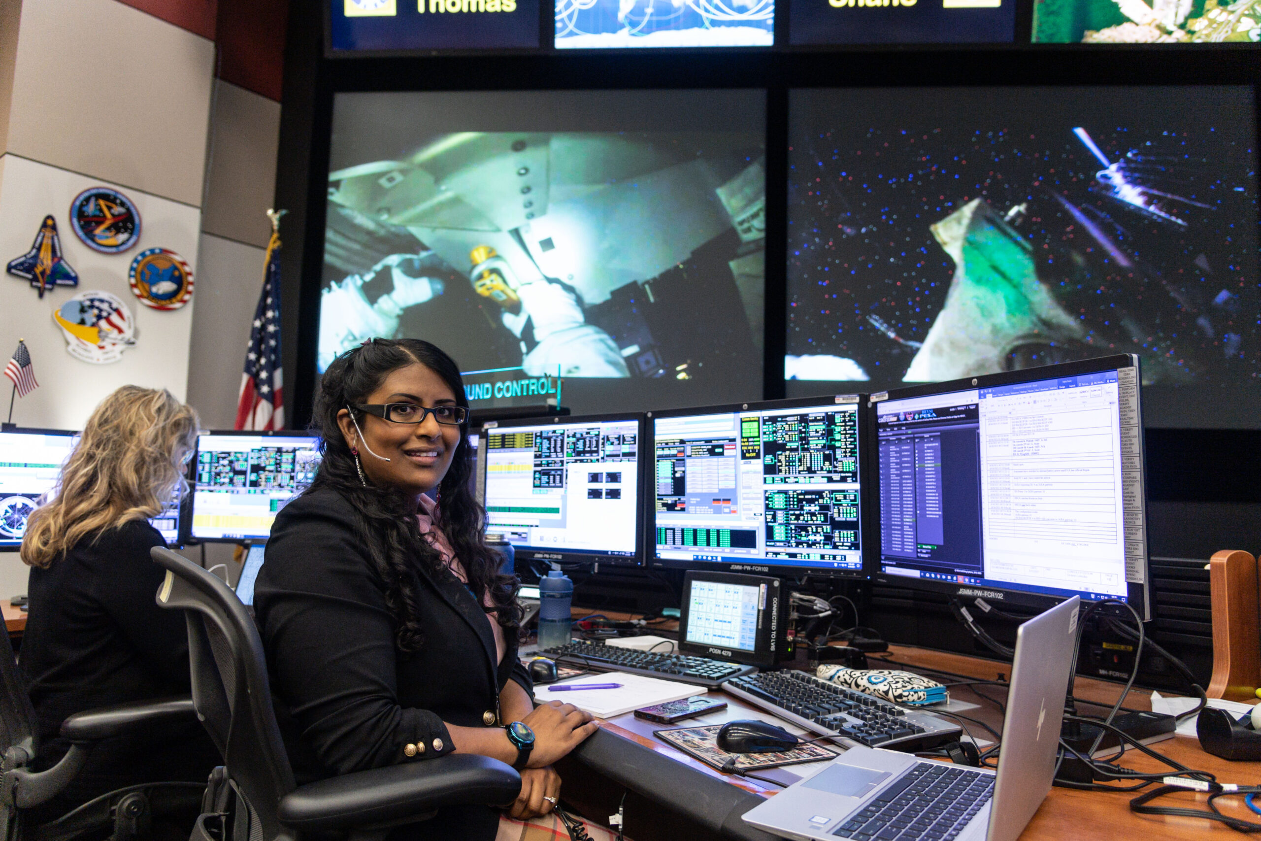 me console2 high.jpg A woman sits at a desk and poses in front of three monitors. Multiple monitors display live data and video of astronauts on the International Space Station. An American flag and NASA mission patches are visible on the wall behind her.