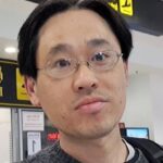 Michiharu Hyogo: Citizen Scientist Leads New NASA Study A man with short black hair, glasses, and a neutral expression is standing in an airport terminal. Yellow signage with directional symbols is visible in the background.