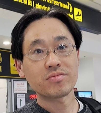 Michiharu Hyogo: Citizen Scientist Leads New NASA Study A man with short black hair, glasses, and a neutral expression is standing in an airport terminal. Yellow signage with directional symbols is visible in the background.