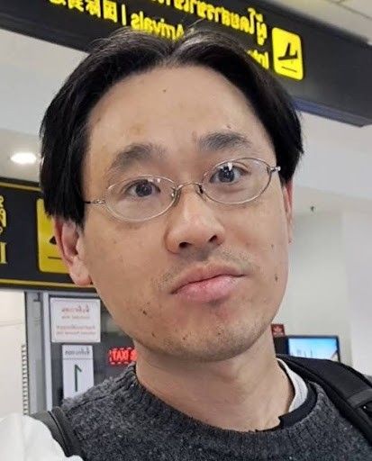 michiharu hyogo.jpg A man with short black hair, glasses, and a neutral expression is standing in an airport terminal. Yellow signage with directional symbols is visible in the background.